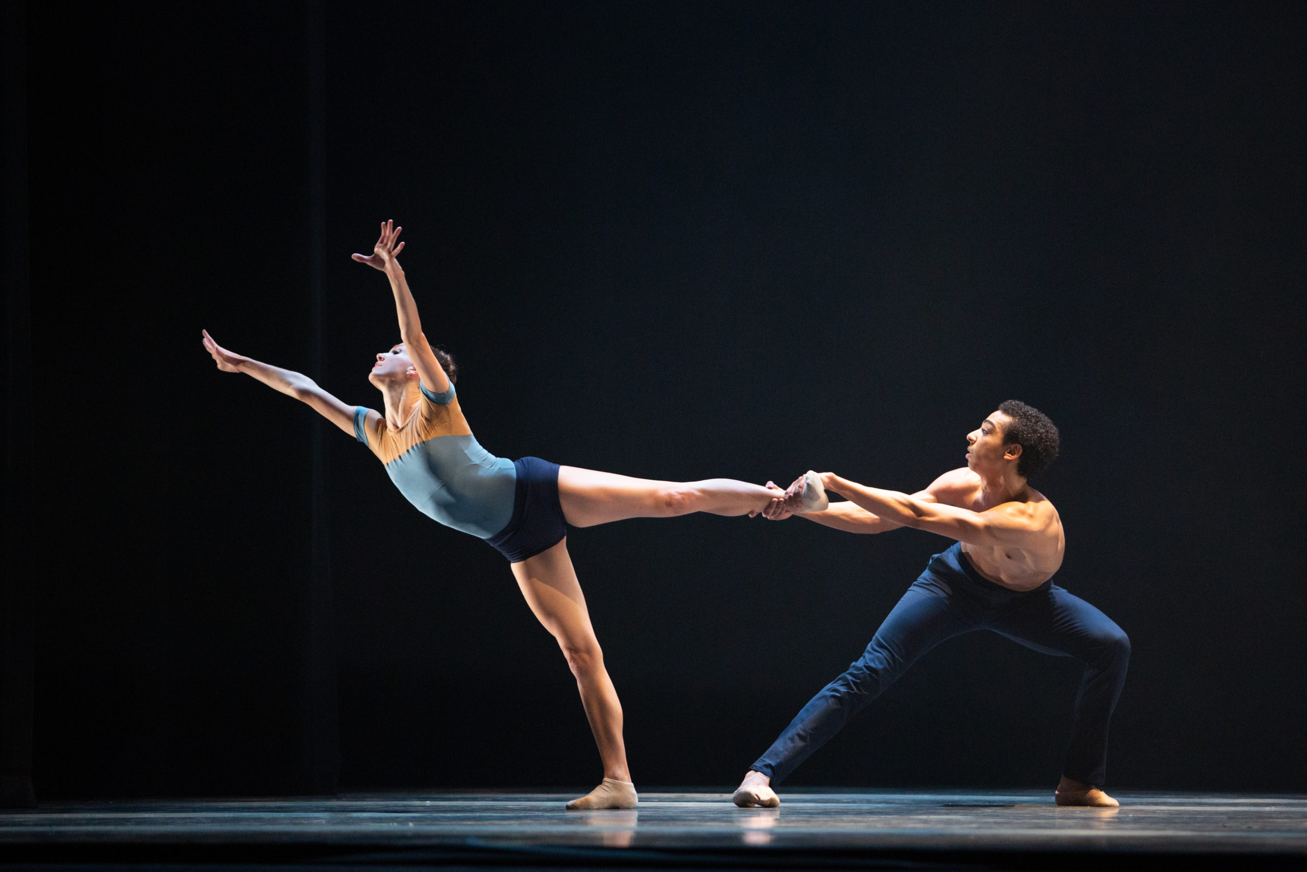 Ethan Price lunges on his left leg and pulls Jillian Barrell's left flexed foot as she stretches in arabesque with her arms reaching out high with fingers splayed. She wears a light blue leotard, dark blue bootie shorts and socks while Price wears dark blue pants and socks.