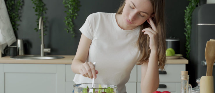 Unhappy woman eating vegetable salad at table in kitchen.