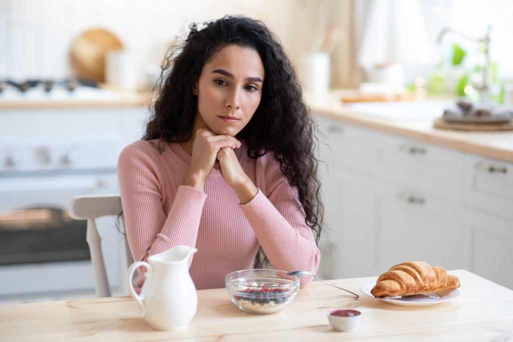 Portrait Of Upset Pensive Woman Sitting At Table In Kitchen With Thoughtful Face Expression
