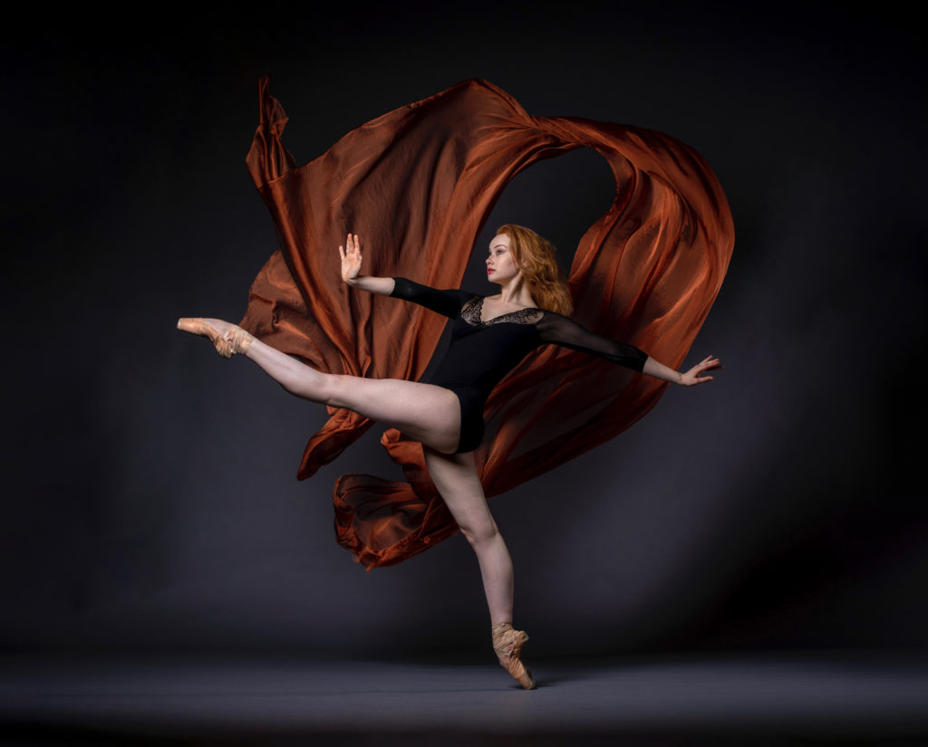 Graceanne Pierce is a fair-skinned woman with red hair. She wears pointe shoes and a black leotard. She faces left, balancing on pointe on her right leg and kicking up her left leg, framed by billowing copper fabric