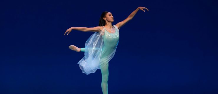 Courtney Holland, costumed in a bright blue unitard under a sheer, loose gown, performs an attitude derriere croisé with her left leg back and her amrs floating out to the side. She dances on a blue-lit stage in front of a blue backdrop.