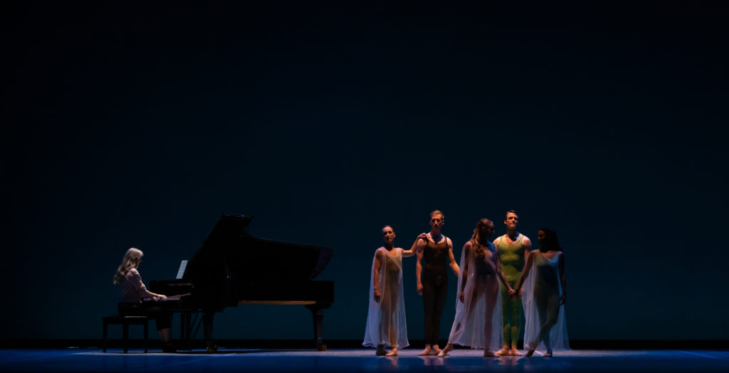 A group of five dancers—three women and two men—stand on stage left in various poses as a pianist performs at a grand piano on stage right. The dancers wear colorful, monochrome unitards, and the women wear a sheer, loose gown on top.