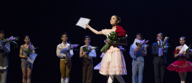 Heesun Kim stands onstage in B plus with her right foot back and holds a bouquet of roses in her left arm. She lifts a paper award up with her right hand and smiles brightly. She wears a pink romantic tutu, pick tights nad pink pointe shoes. Behind her, male and female dancers in various costumes stand in a line and applaud.