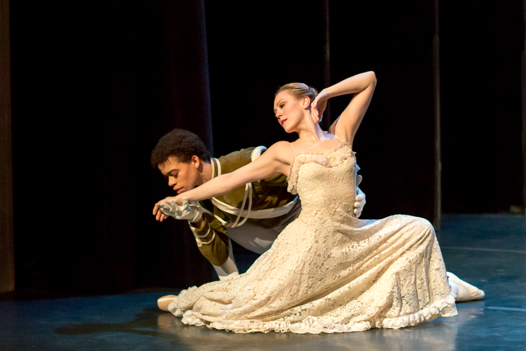 During a performance, Elena Zahlmann, wearing a long, lacy off-white dress, lunges onto her right knee and looks back, extending her right hand to Steven Melendez as he crouches behind her and kisses it. Melendez wears a green military-style jacket and white tights and shoes.