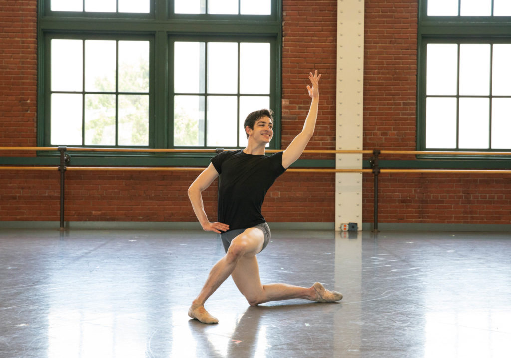 Joshua Kiesel, in a Black T-shirt and gray bike shorts,poses on his right knee in a large dance studio with brick walls and large windows. He places his right arm on his hip and lifts his left arm up above his head, and looks toward the left corner with a large smile.