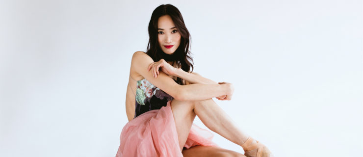 Fumi Kaneko sits on the floor on a white backdrop and crosses her right leg over her left knee, pointing her foot and curling her arms around her right knee. Her hair is long around her shoulders and she wears a dark floral leotard and long, voluminous and gauzy pink skirt. She smiles confidently with a closed mouth and looks directly at the camera.