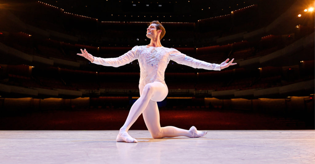 Jacopo Tissi kneels onstage with his back facing the house. He kneels on his right leg and extends his arms out, palms facing up, and looks off to the right with a large smile. He wears a white tunic, white tights and white ballet slippers.