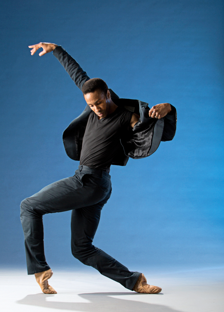 Jermel Johnson is in a parallel fourth position on forced arch. He is wearing navy blue pants, a matching blazer, a black t-shirt and flesh-toned ballet shoes. He is opening the upper left side of his blazer as he twists his torso toward the camera.