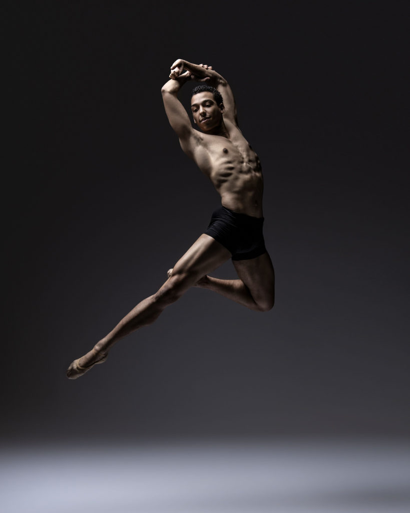 Ethan Price, wearing black shorts and brown ballet slippers, jumps high with his right leg extended long and slightly out to the side and his left leg in passé. His arms are curved above his head and he grasps his left arm with his right hand.