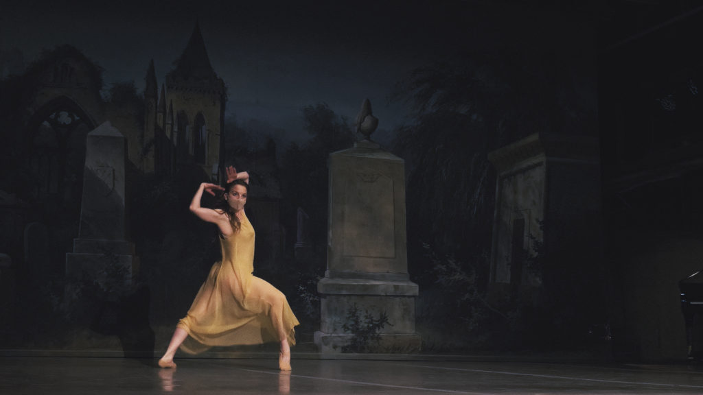 Jaime Lynn WItts, wearing a filmy yellow dress and face mask, dances in front of a backdrop showing a cemetary at night. She lunges forward on her right leg and pops her right foot up on demi-pointe, and lifts her bent arms to encircle her head.