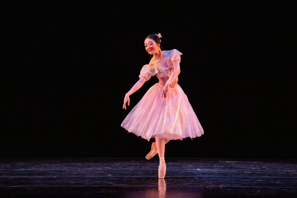 Wearing a pink Romantic tutu with puffed short sleeves, Mayu Odaka performs onstage in rfont of a black backdrop. She poses on pointe in a coupé derriere with her left foot back. Her arms sweep across her body towards the right.