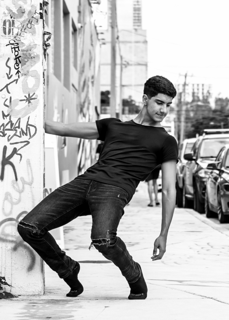 With his right hand, Tristan Barron holds on to a graffitied wall on a city sidewalk and lunges forward in parallel onto a high demi-pointe. He wears a black T-shirt and jeans, and black ballet slippers, and looks down towards his left arm which hangs down.
