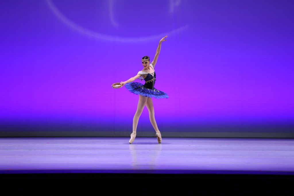 Siena España, wearing a blue tutu with white short sleeves and a gold headband across her forehead, poses in fourth position effacé on pointe facing stage lwft. She lifts her left arm up high and her right arm out to the side, and holds a tambourine in her right hand.