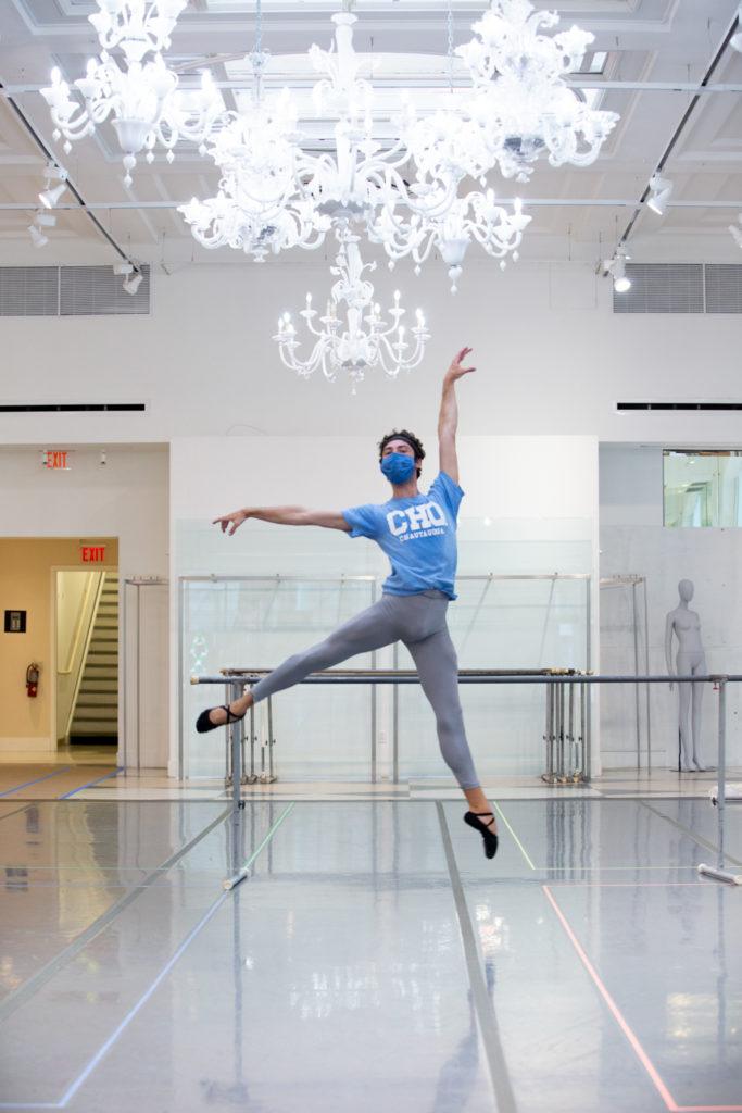 In a brightly lit studio with portable ballet barres and an ornate chandelier, dancer Ben Lepson performs a temps levé in first arabesque with his right leg back. He wears a blue T-shirt and face mask, gray tights and black ballet slippers.