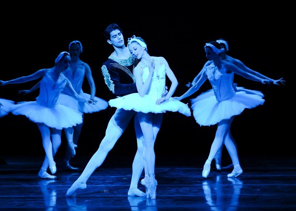 Alexandra Kochis and Alejandro Diaz dance as Odette and Siegfried. Alexandra wears a white tutu and a feather crown headpiece. She stands in 5th on pointe gazing down to the side as Alejandro, wearing white tights and a black, long-sleeve top, stands behind her and holds her waist and looks to her head, his right leg in tendu to the side.