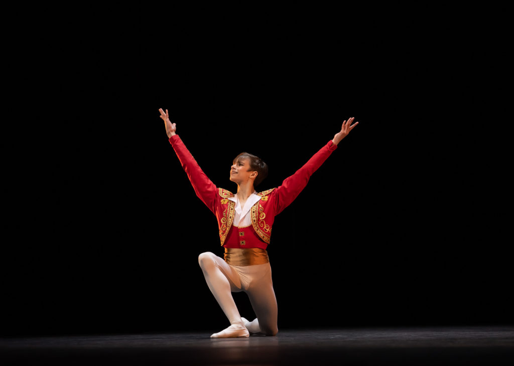 Alexei Orohovsky kneels on his left knee as he finishes a dance onstage. He lifts his arms high in a wide V-shape and smiles confidently towards the audince. He wears a birght red bolero jacket and gold cumberbund, white tights, and white ballet slippers, and poses in front of a black backdrop.