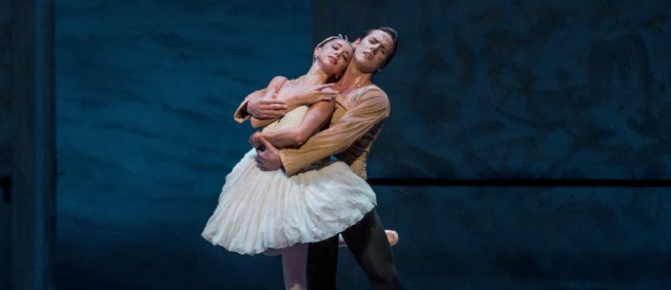 April Daly wears a white tutu and leans back onto her male partner. Her partner's arms wrap around her from behind, and her arms cross in front of her. She stands on pointe on her left leg with her right leg wrapped behind her partner.