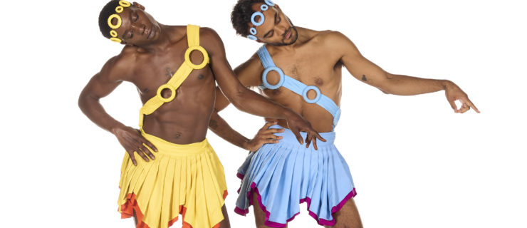 Cemiyon Barber and Taylor Stanley pose barefoot in front of a white background. They wear matching costumes, Barber in yellow and orange and Stanley in blue and pink. Their hoop headbands and pleated skirts are identical other than the color. Their sashes with hoops cross from shoulder to hip in opposite directions, and Stanley's left ankle has a hoop anklet while Barber's is on his right. Their right hands are on their hips, slightly jutted out to the side. They incline their heads to the right looking at their outstretched winged left feet. Their left arms are held loosely to the side with fingers pointing down.