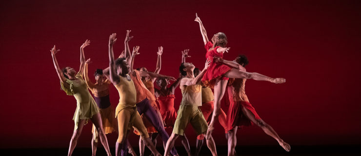 A large group if dancers from Dance Theatre of Harlem stand close together onstage with their legs in a wide stance facing stage left. They lift their arms up above their heads and arch slightly back in reaction to the dancer coming towards them. A female dancer in a red dance dress performs as temps levé towards the group as two men lift her by the waist. The dancers wear multi-colored costumes and perform in front of a red backdrop.