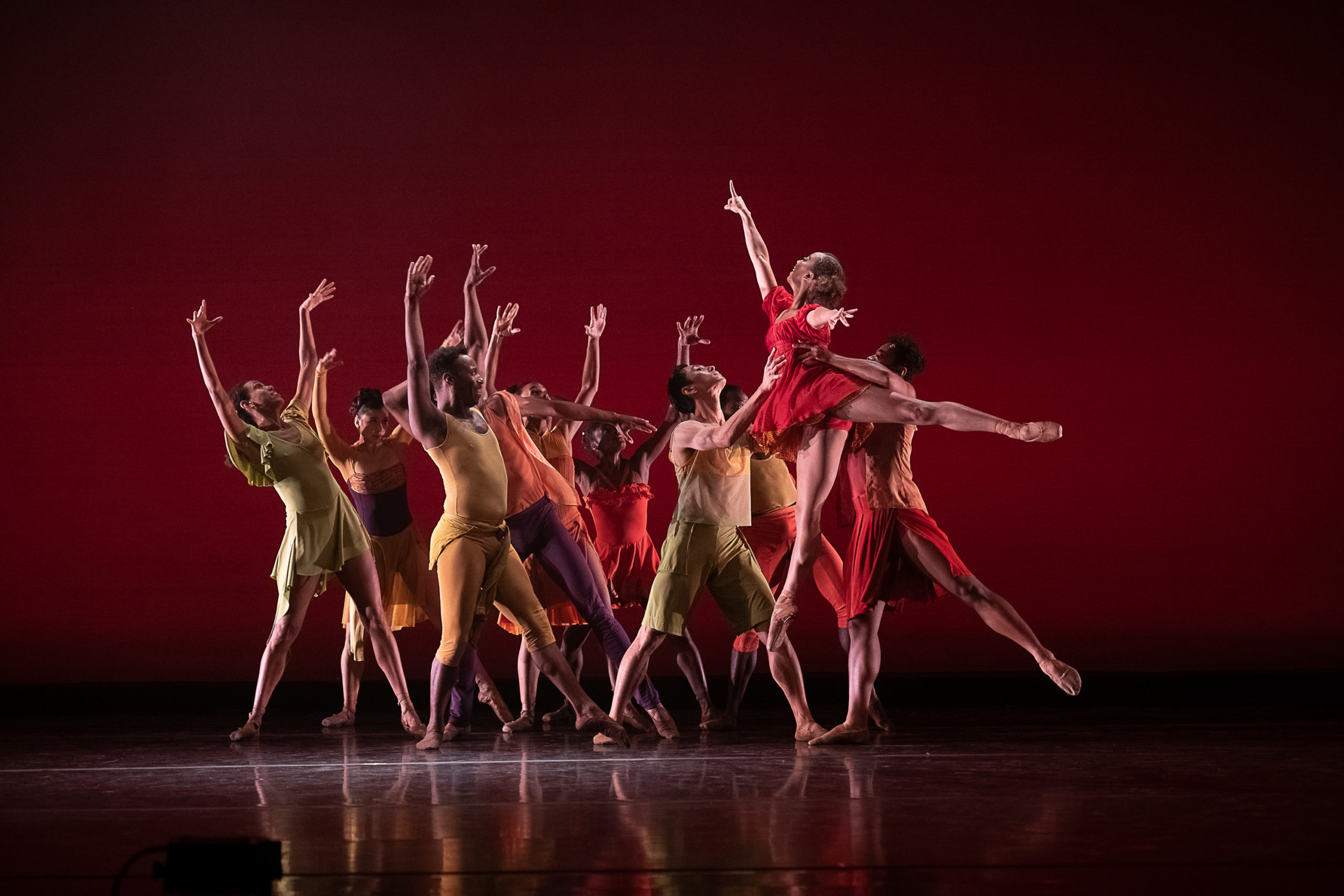 A large group if dancers from Dance Theatre of Harlem stand close together onstage with their legs in a wide stance facing stage left. They lift their arms up above their heads and arch slightly back in reaction to the dancer coming towards them. A female dancer in a red dance dress performs as temps levé towards the group as two men lift her by the waist. The dancers wear multi-colored costumes and perform in front of a red backdrop.
