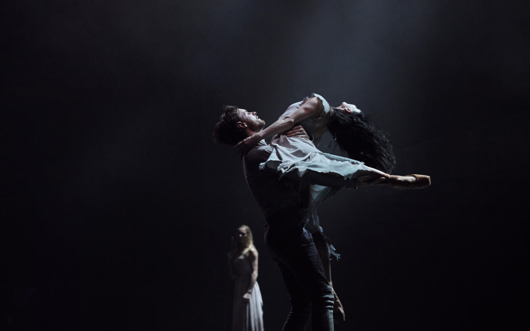 James Streeter lifts Tamara Rojo with his left arm wrapped around her back and his right arm lifting her back leg. She arches back in a dramatic arabesque with her stomach on his chest and long dark hair flying back. In the background, a female dancer stands and watches, illuminated against a dark backdrop.