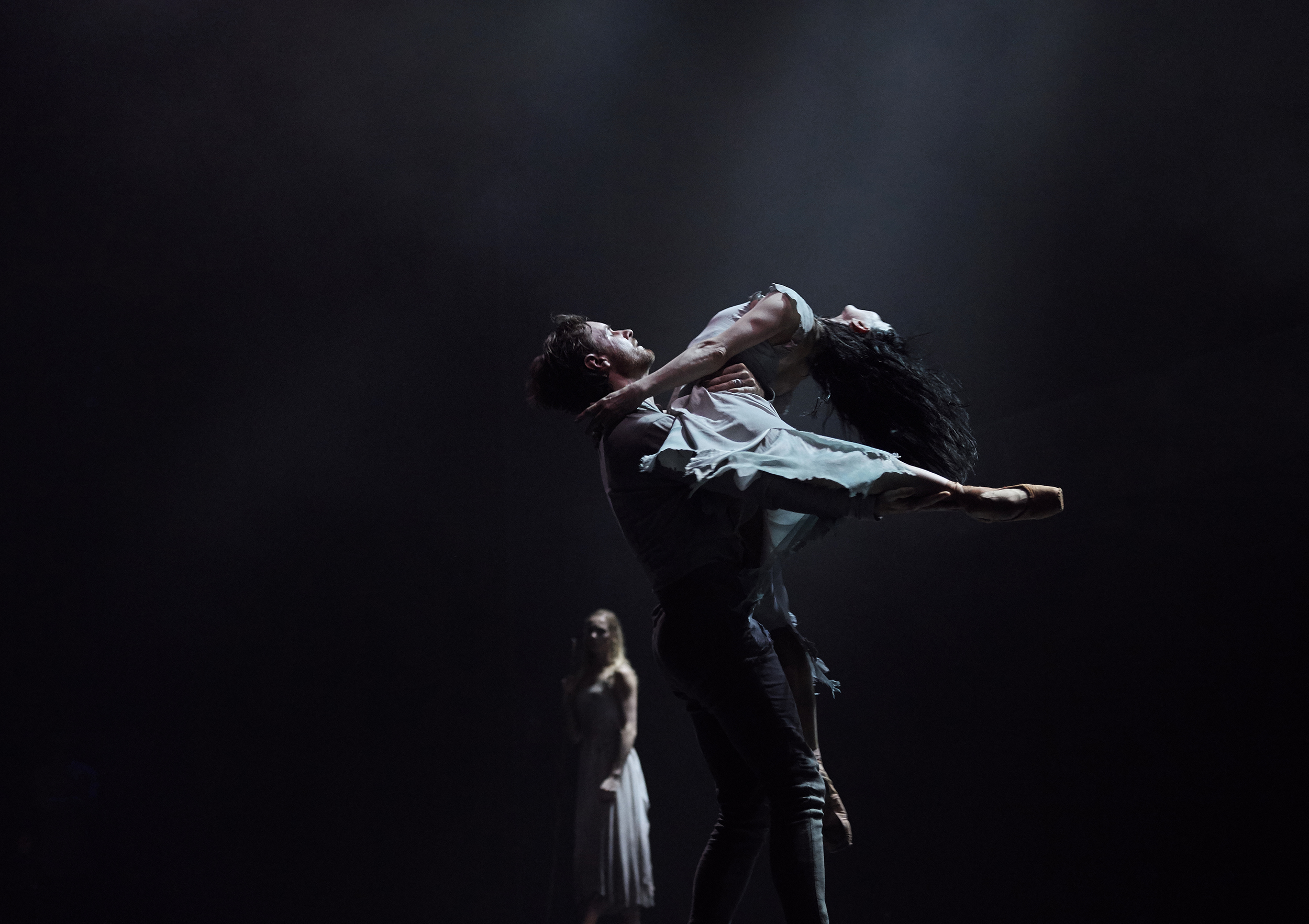 James Streeter lifts Tamara Rojo with his left arm wrapped around her back and his right arm lifting her back leg. She arches back in a dramatic arabesque with her stomach on his chest and long dark hair flying back. In the background, a female dancer stands and watches, illuminated against a dark backdrop.