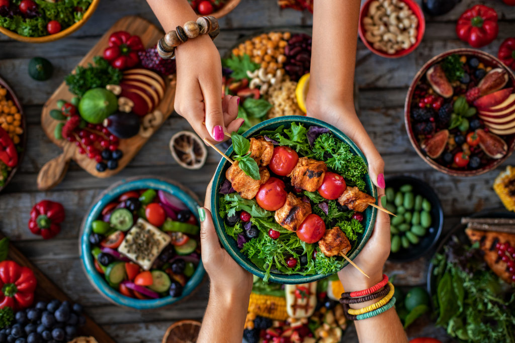 An overhead shot of two people passing a bowl of greens and chicken skewers above a full spread of colorful food.