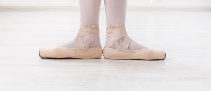 A ballerina is shown from the calves down in first position. She wears pink tights and pink pointe shoes and stands on a white floor in front of white walls.