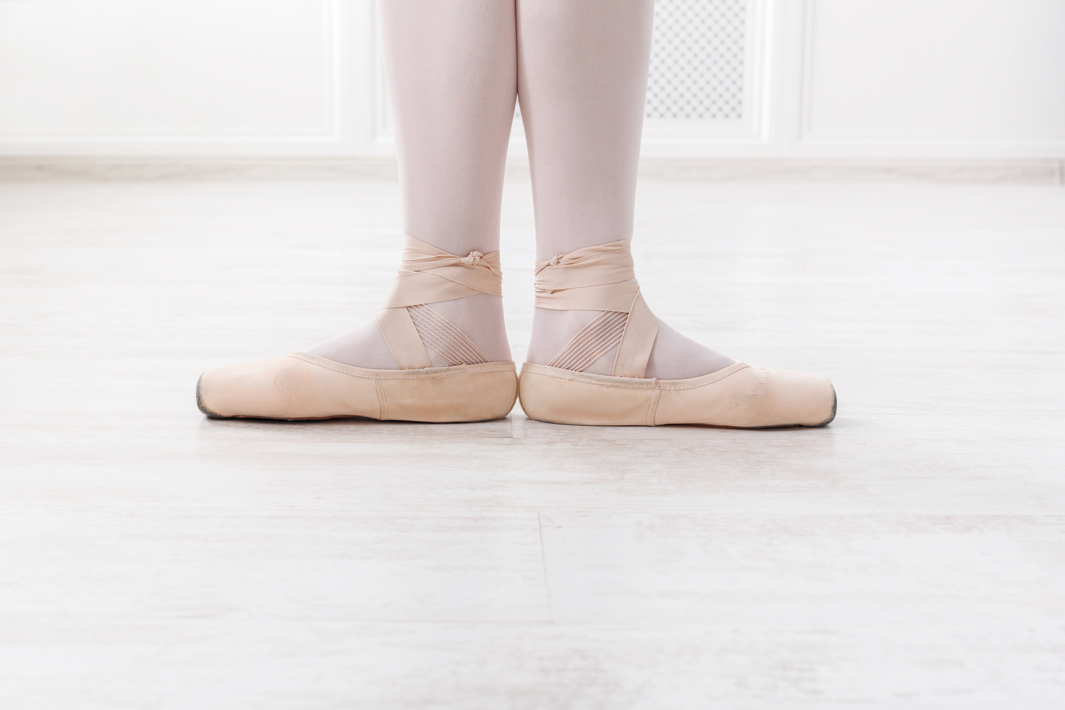 A ballerina is shown from the calves down in first position. She wears pink tights and pink pointe shoes and stands on a white floor in front of white walls.