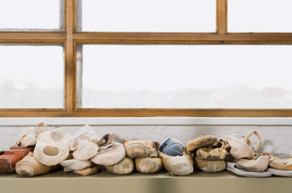 A pile of used pink and tan pointe shoes and ballet flat shoes rests on a tan shelf in front of a white wall and brightly lit window with wooden rails.