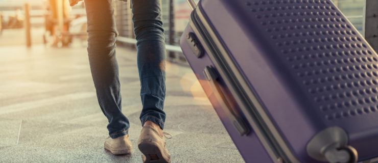 A female in jeans, a tan shirt and worn white shoes (shown legs-down) pulls a large purple suitcase behind her as she walks through an airport terminal.