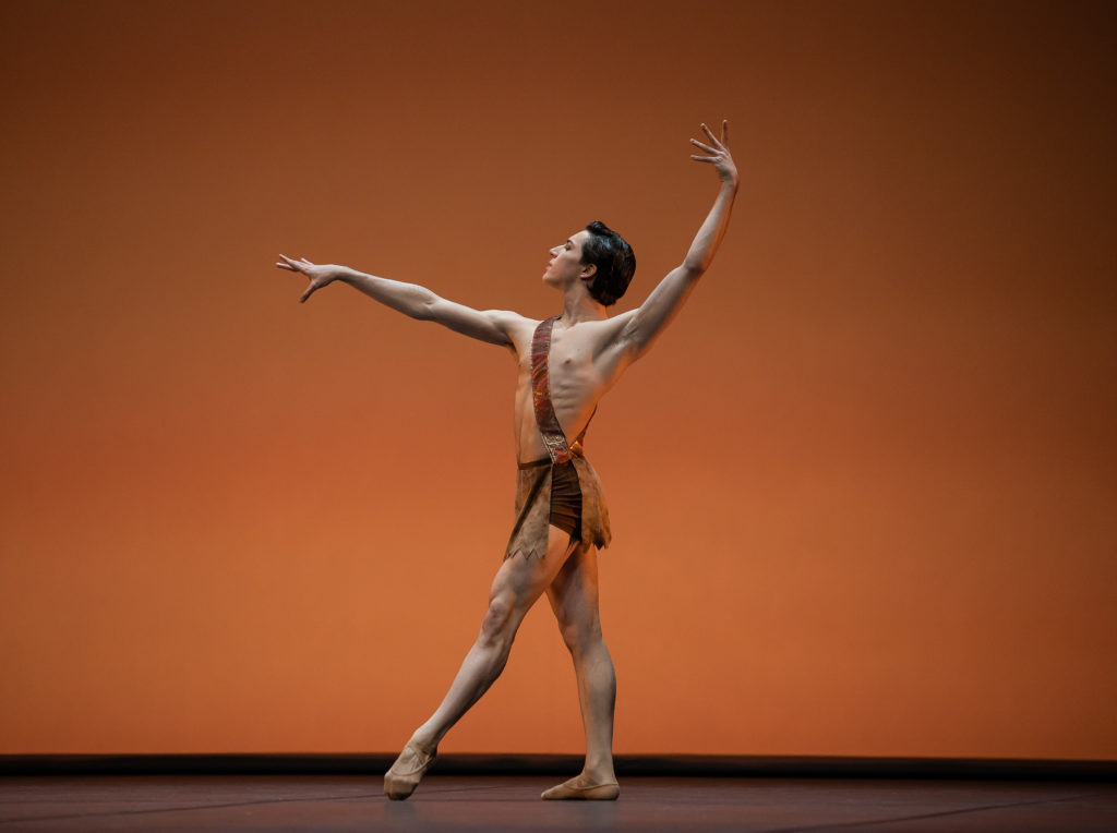 Giulio Diligente stands onstage in front of a bronze-lit backdrop, posing in a tendu devant croisé with his left leg in front. He holds his right arm outstretched in front of him and his left arm up, and wears a brown loincloth costume with a strap around his right shoulder, and brown ballet slippers. He looks out over his right hand confidently.