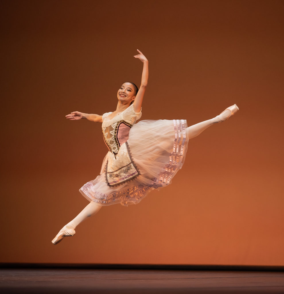 Hui Wen Peng wears a pink peasant dress costume and performs a large sissone fermé onstage in front of a bronze-lit backdrop. She holds her right arm out to the side and her left arm up She wears pink tights and pointe shoes, and looks out towards the audience with a happy smile.