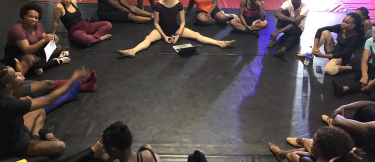 Members of Global Ballet Teachers sit in a circle on the floor of a dance studio dressed in various dancewear garments. GBT co-founder Cecilia Iliesiu sits in a relaxed middle split, holding her hands on the floor and listens with a gentle smile, head turned toward the speaker on the left side of the circle.