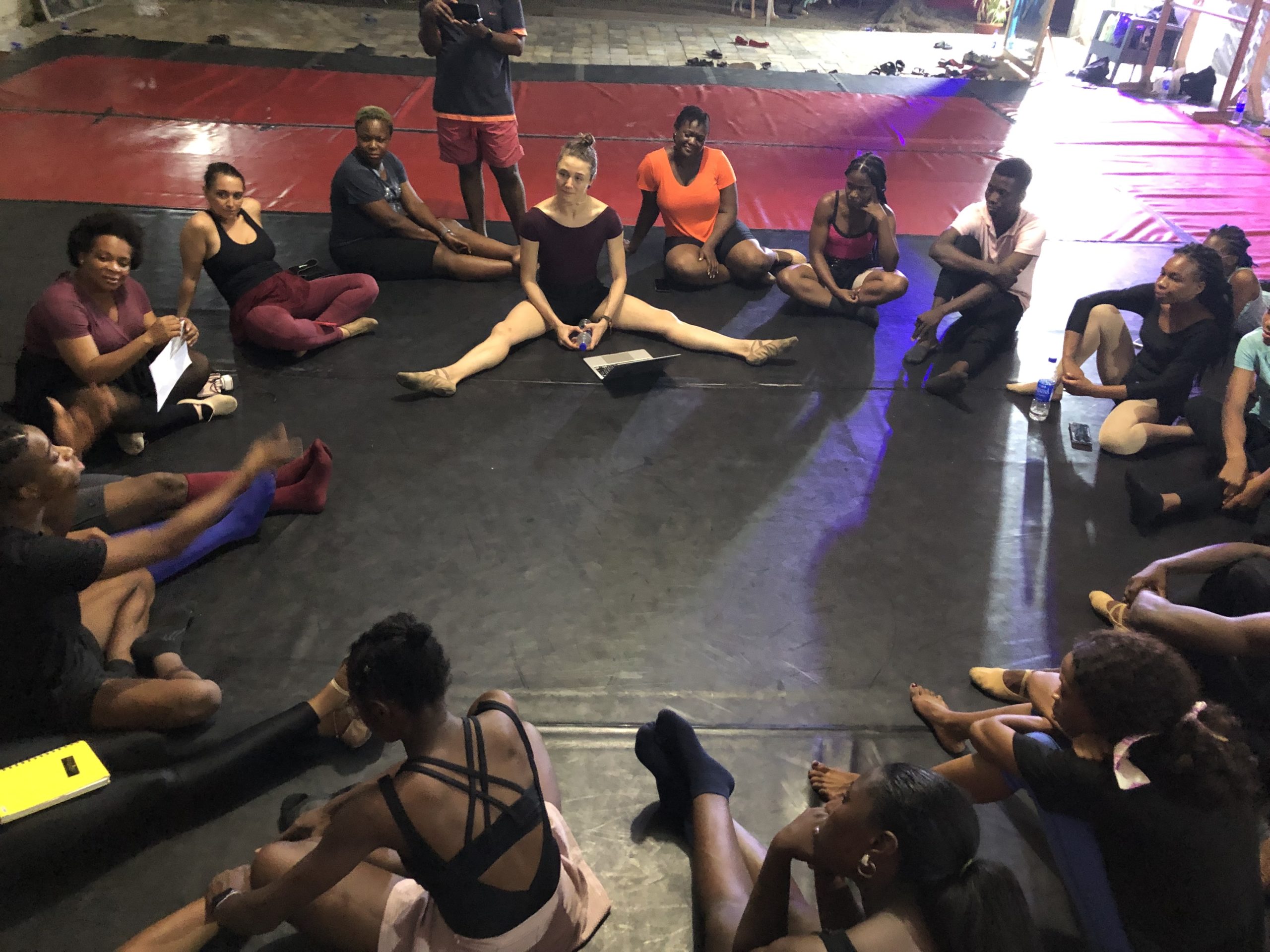 Members of Global Ballet Teachers sit in a circle on the floor of a dance studio dressed in various dancewear garments. GBT co-founder Cecilia Iliesiu sits in a relaxed middle split, holding her hands on the floor and listens with a gentle smile, head turned toward the speaker on the left side of the circle.