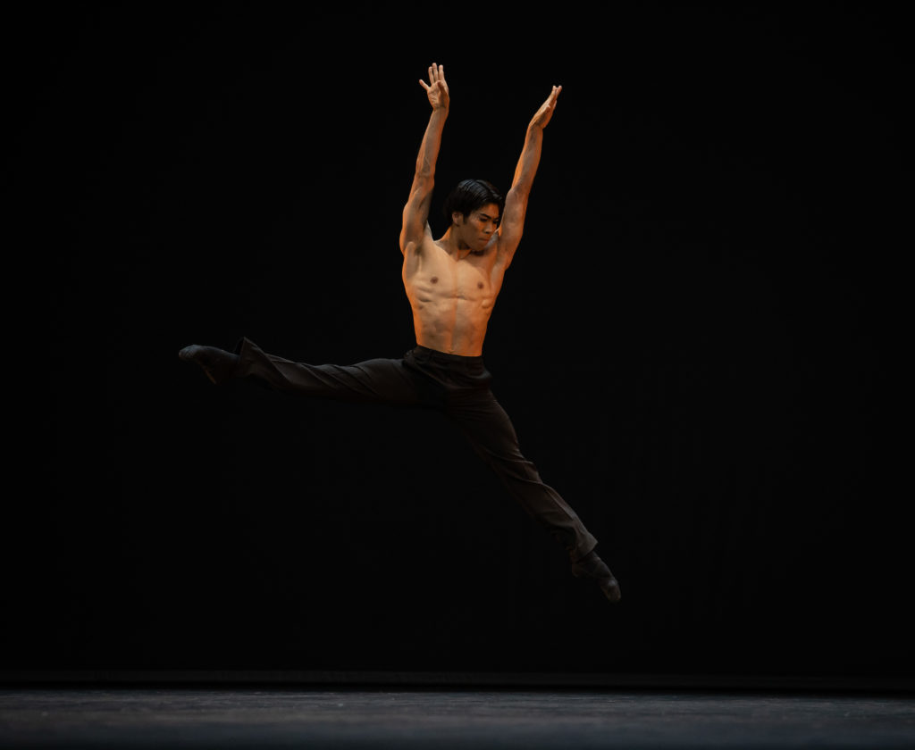 Keita Fujishima performs a sissone onstage moving towards stage left. Shirtless, he wears black pants, socks and ballet slippers and stretches his arms high above his head. He turns his head and looks down towards his left.
