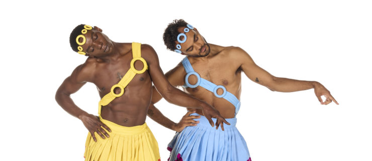 Cemiyon Barber and Taylor Stanley pose barefoot in front of a white background. They wear matching costumes, Barber in yellow and orange and Stanley in blue and pink. Their hoop headbands and pleated skirts are identical other than the color. Their sashes with hoops cross from shoulder to hip in opposite directions, and Stanley's left ankle has a hoop anklet while Barber's is on his right. Their right hands are on their hips, slightly jutted out to the side. They incline their heads to the right looking at their outstretched winged left feet. Their left arms are held loosely to the side with fingers pointing down.