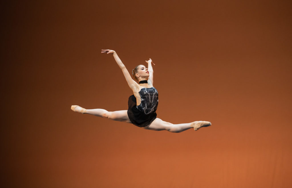 Rinja Rissanen performs a saut de chat onstage towards stage left during a performance, in front of a bronze-lit backdrop. She wears a short black skirt and patterened black and gray bodice, with a black choker around her neck, and tan pointe shoes. She lifts her arms high and slightly behind her as she jumps.