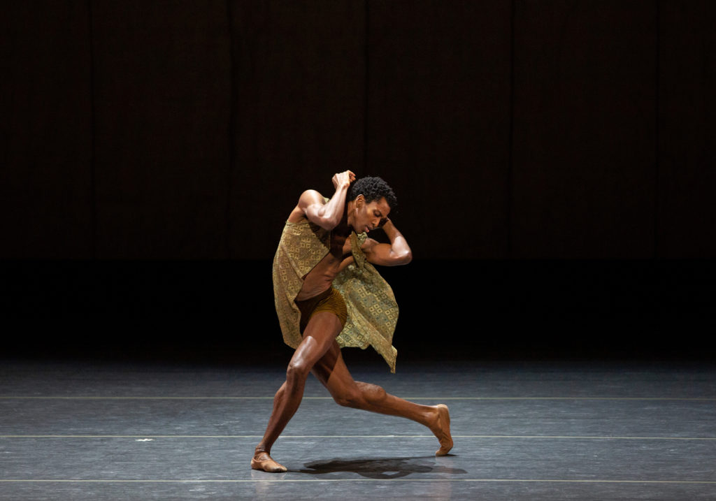 Calvin Royal III dances onstage. He is wearing a patterned, dark green, loosely fitting top and dark green shorts.