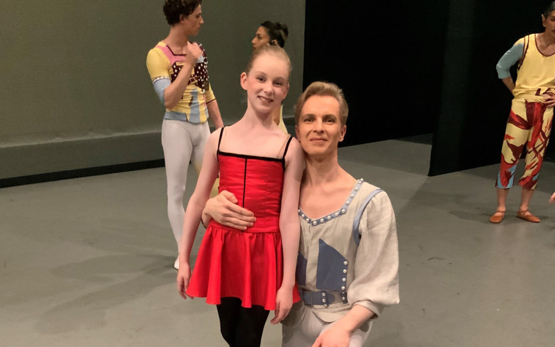 Tiit Helemits and his daughter Chloe pose backstage before a performance. Tiit, who is wearing a gray tunic with blue trim and gray tights, kneels down next to his daughter, who wears a bright red dress with black spaghetti straps and black tights. They smile for the camera. Behind them, two dancers in multicolored costumes chat.