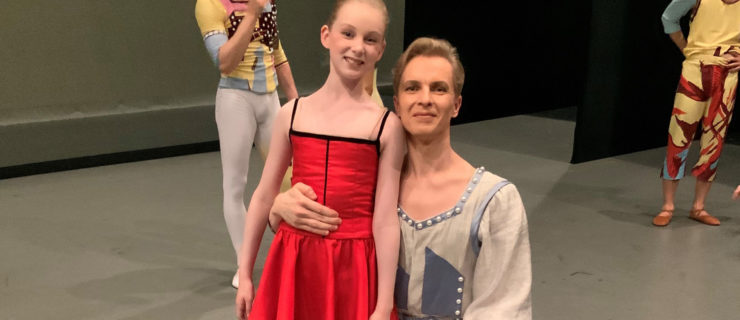 Tiit Helemits and his daughter Chloe pose backstage before a performance. Tiit, who is wearing a gray tunic with blue trim and gray tights, kneels down next to his daughter, who wears a bright red dress with black spaghetti straps and black tights. They smile for the camera. Behind them, two dancers in multicolored costumes chat.