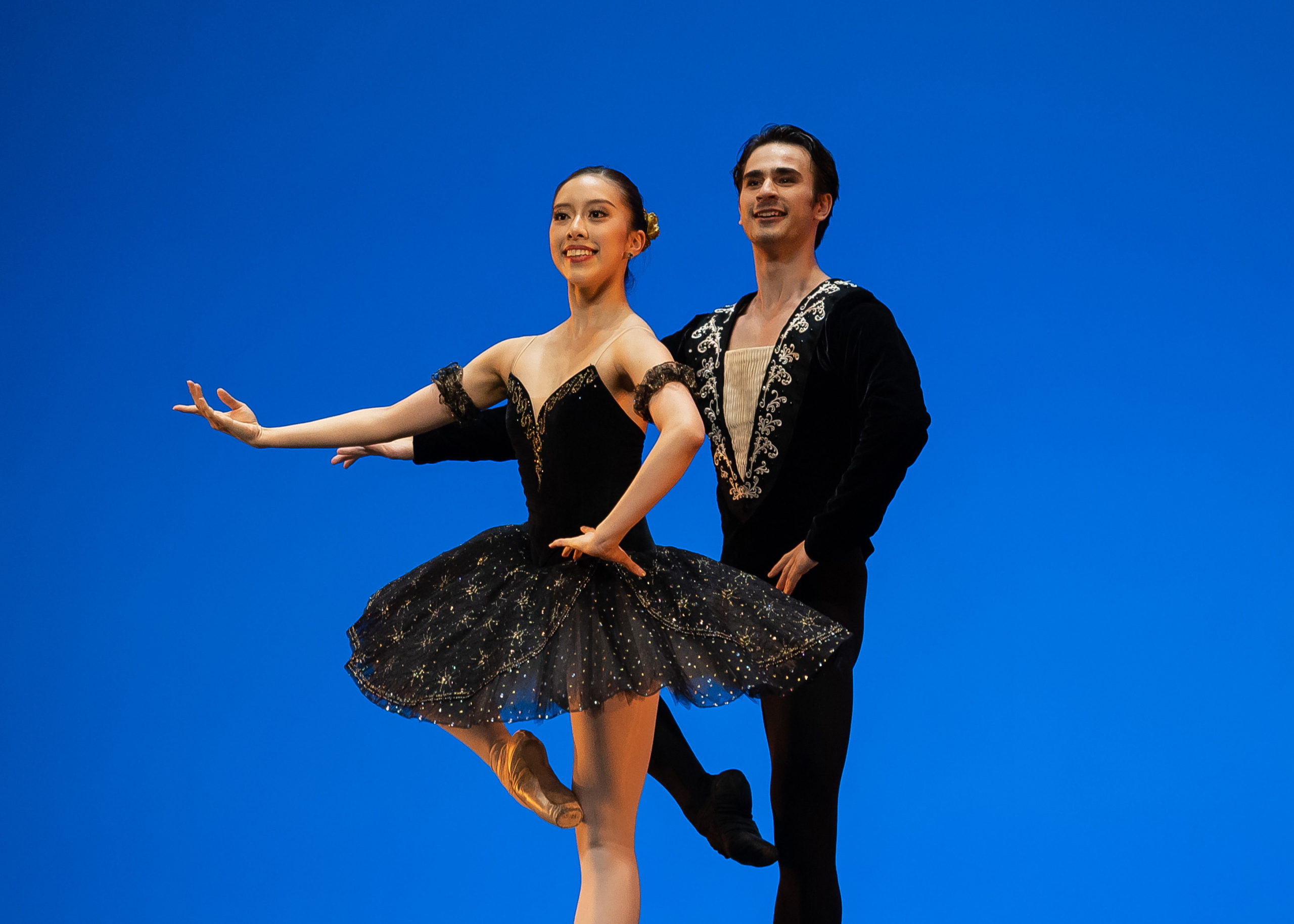 Yuka Masumoto, in a black tutu, and Clark Eselgroth, in a black tunic and tights, are shown from the knee up performing onstage in front of a blue backdrop. Masumoto stands in front of Eselgroth, and they both perform a passé with their right leg and hold their right arm out with an open palm. Their left hands are placed on their hips, and they both smile towards the audience.