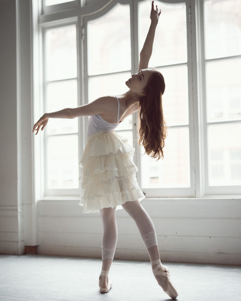 Alexandra Martin stands in profile in tendu derriere with her left leg back and her left arm in front of her. She arches back, lifting her right arm high, and her long hair flows down her back. She wears a white leotard, fluffy off-white skirt, pink tights and pink pointe shoes.