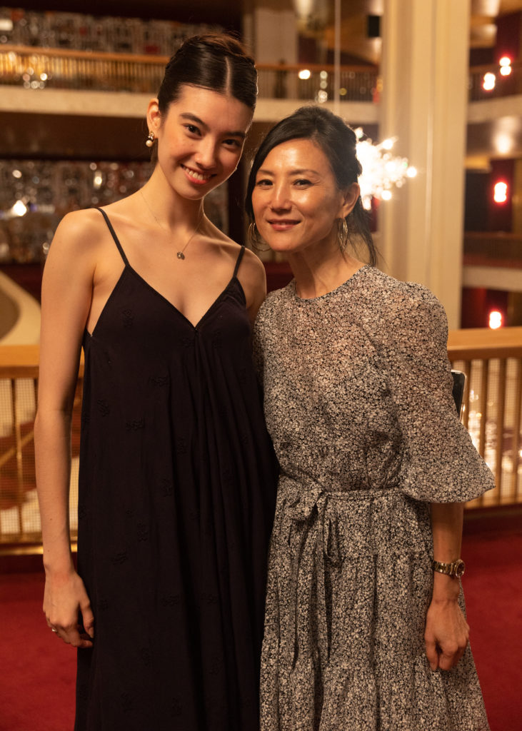 CHloe MIsseldine, in a long, black spaghetti-strap dress, stands next to her mom Yan Chen, who wears a gray-patterned long-sleeved dress. They wrap their arms around each other's waists and smile towards the camera. They stand in the mezzanine of a large opera house, with a mid-century modern crystal chandelier in the background.