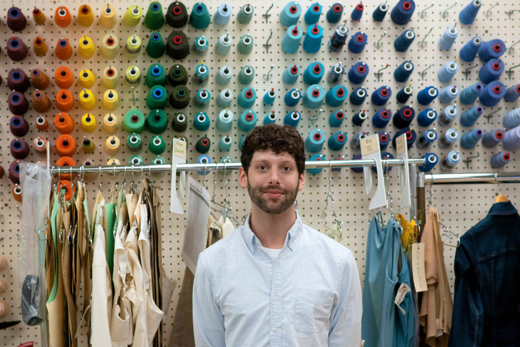 Lovesky wears a collared shirt, standing in front of a wall filled with thread and racks of clothing. He is looking directly at the camera and softly smiling.