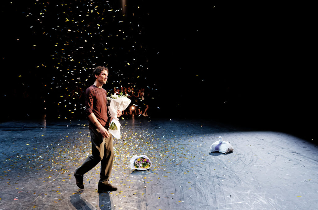 Stéphane Bullion, dressed in a brown shirt and brown pants and shoes, holds a bouquet of flowers in his left amr and walks forward on stage. Confetti rains down on him from above, and other bouquets of flowers litter the stage.