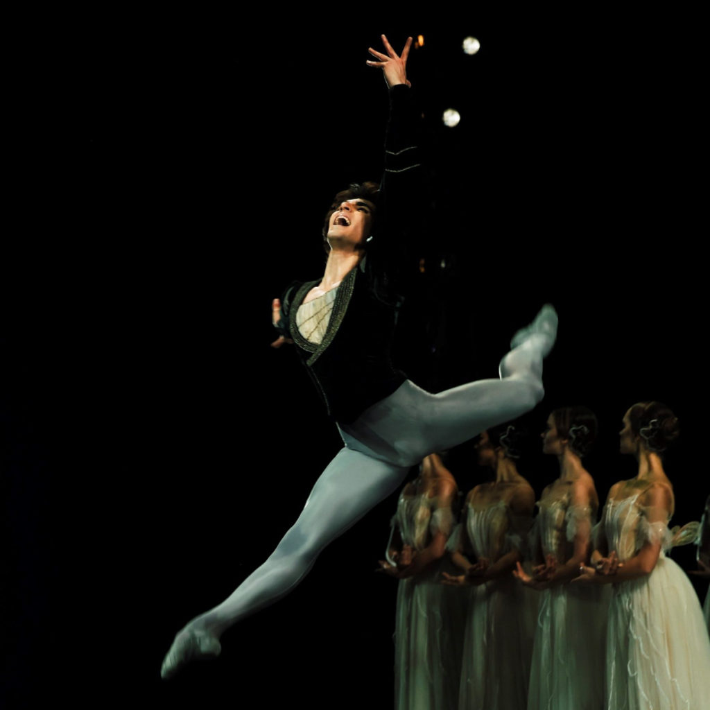 Wearing gray tights and ballet slippers and a dark, velvet tunic, Cesar Corrales performs an explosive sissone in attitude croisé with his left leg behind him. He reaches for the sky with his left hand and looks up with a look of anguish. Behind him onstage, a line of four women in long white tutus stand in B plus with their lower arms crossed at their waist, and look towards the wings on stage right.