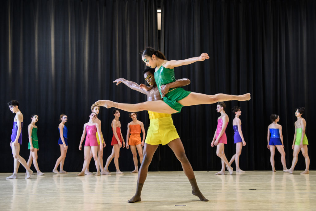During an in-studio dress rehearsal, a young male and female dancer partner together. He stands with his legs apart and holds the female by the waist with his left arm as she performs a grand jeté with her right arm around his neck and left arm out to the side. He wears bright yellow shorts and brown socks; she wears a green bodice and shorts and tan socks.