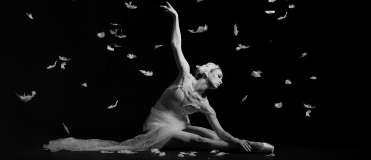 In this black and white photo, Megan Jacobs sits on the ground with her right leg extended in front of her and her left leg tucked. She leans over her legs, touching her left hand to her right foot, and reaches her right arm back behind her. She wears a feathered headpiece, pointe shoes, a light-colored leotard and a swath of white tulle wrapped around her waist, which trails out onto the ground. Feathers rain down softly from the ceiling.