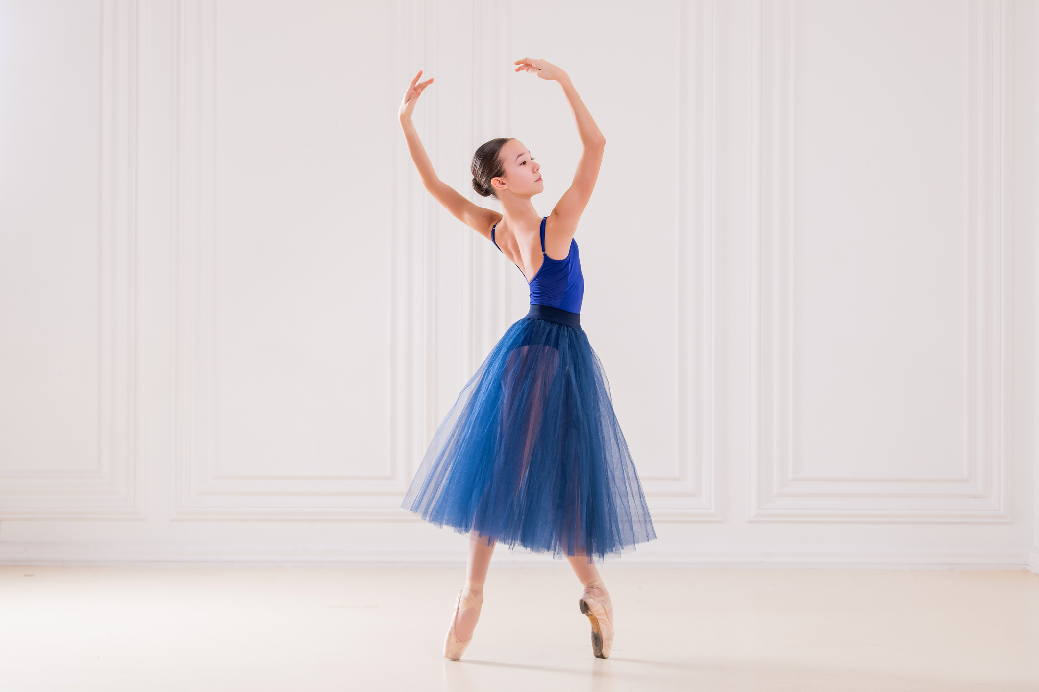 Ask Amy. A young dancer in fourth position on pointe with her arms in fifth position and her torso twisting to the left. She is wearing a blue leotard and romantic length tutu and dancing in a white-walled studio.